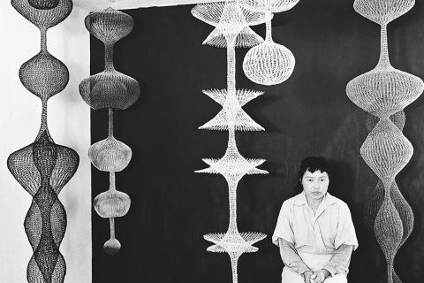 Black and white photograph of artist Ruth Asawa surrounded by her wire mesh sculptures that hang from the ceiling.